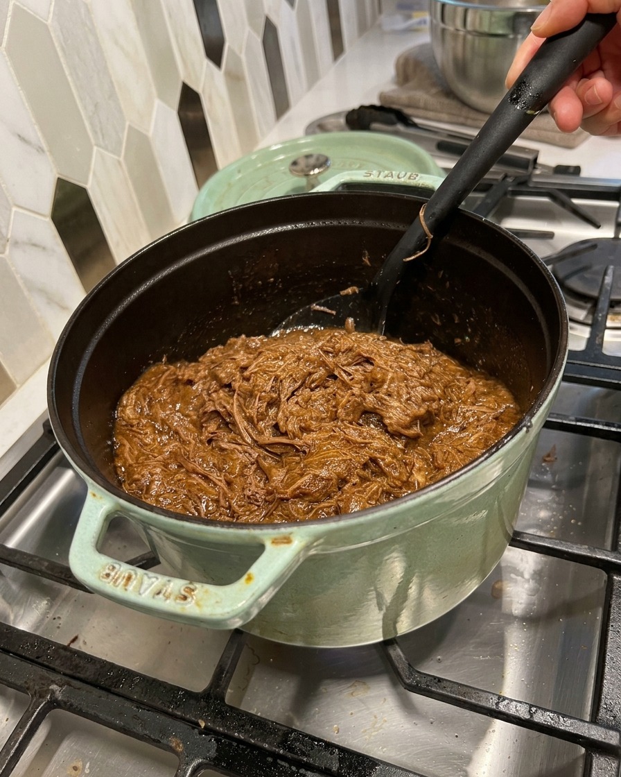 Bœuf effiloché mélangé à la sauce barbecue avec une spatule noire dans la cocotte.