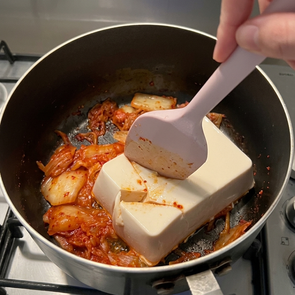 Une spatule en silicone rose coupant un bloc de tofu soyeux blanc sur du kimchi rouge dans une casserole.