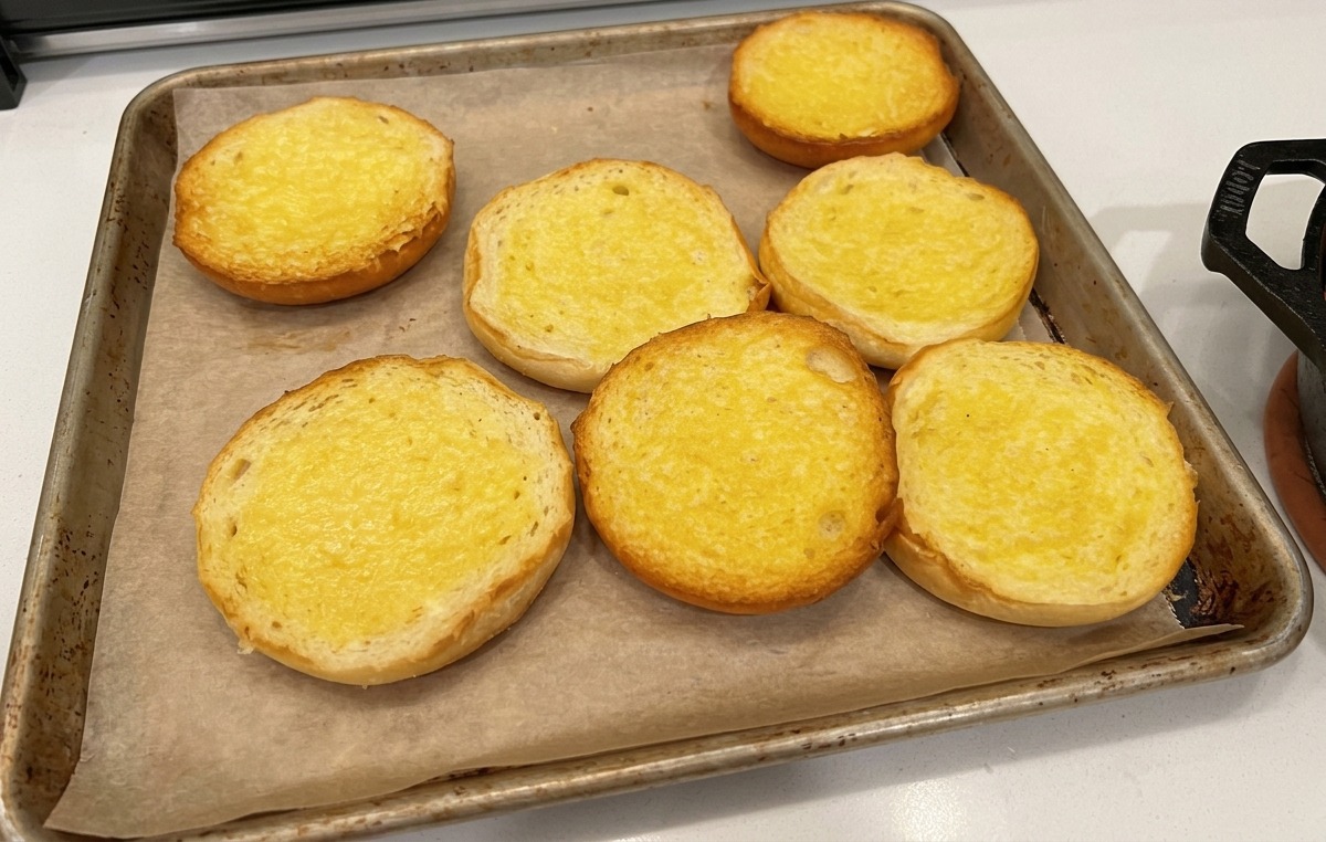 Moitiés de pains à burger beurrées disposées sur du papier sulfurisé sur une plaque de cuisson.