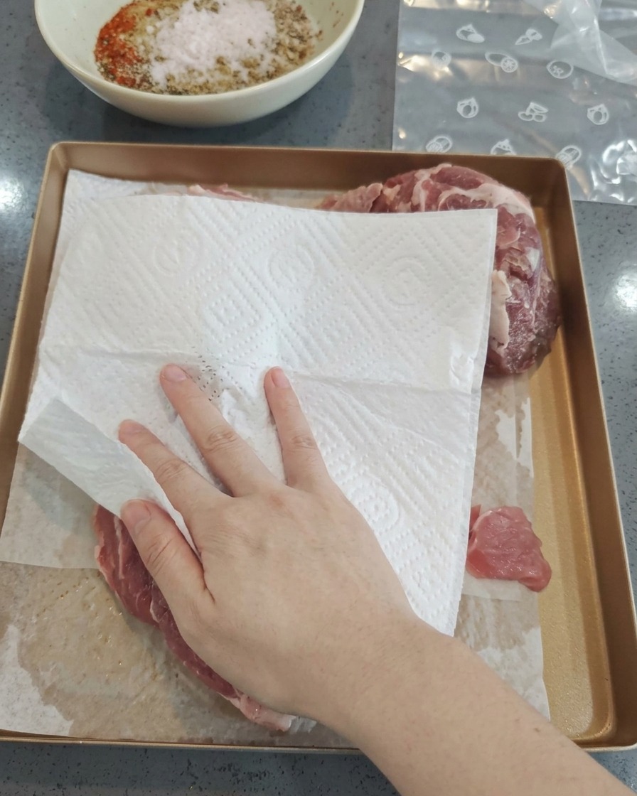 Une main utilisant un essuie-tout pour éponger des morceaux de porc cru sur une plaque.
