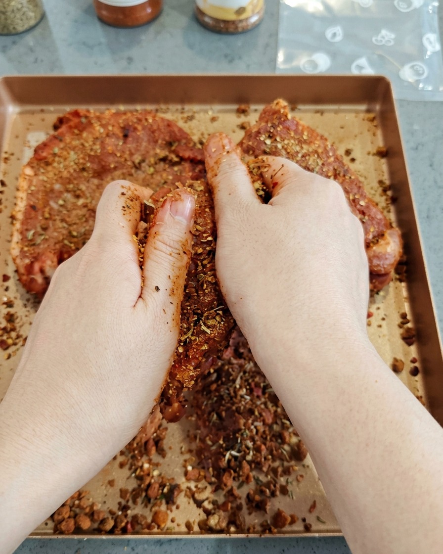 Des mains frottant vigoureusement un mélange dépices sombre sur de gros morceaux de porc cru sur une plaque.