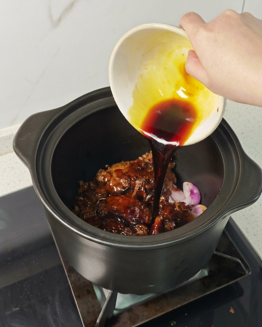 Une main versant une sauce sombre depuis un petit bol sur le porc dans la cocotte.