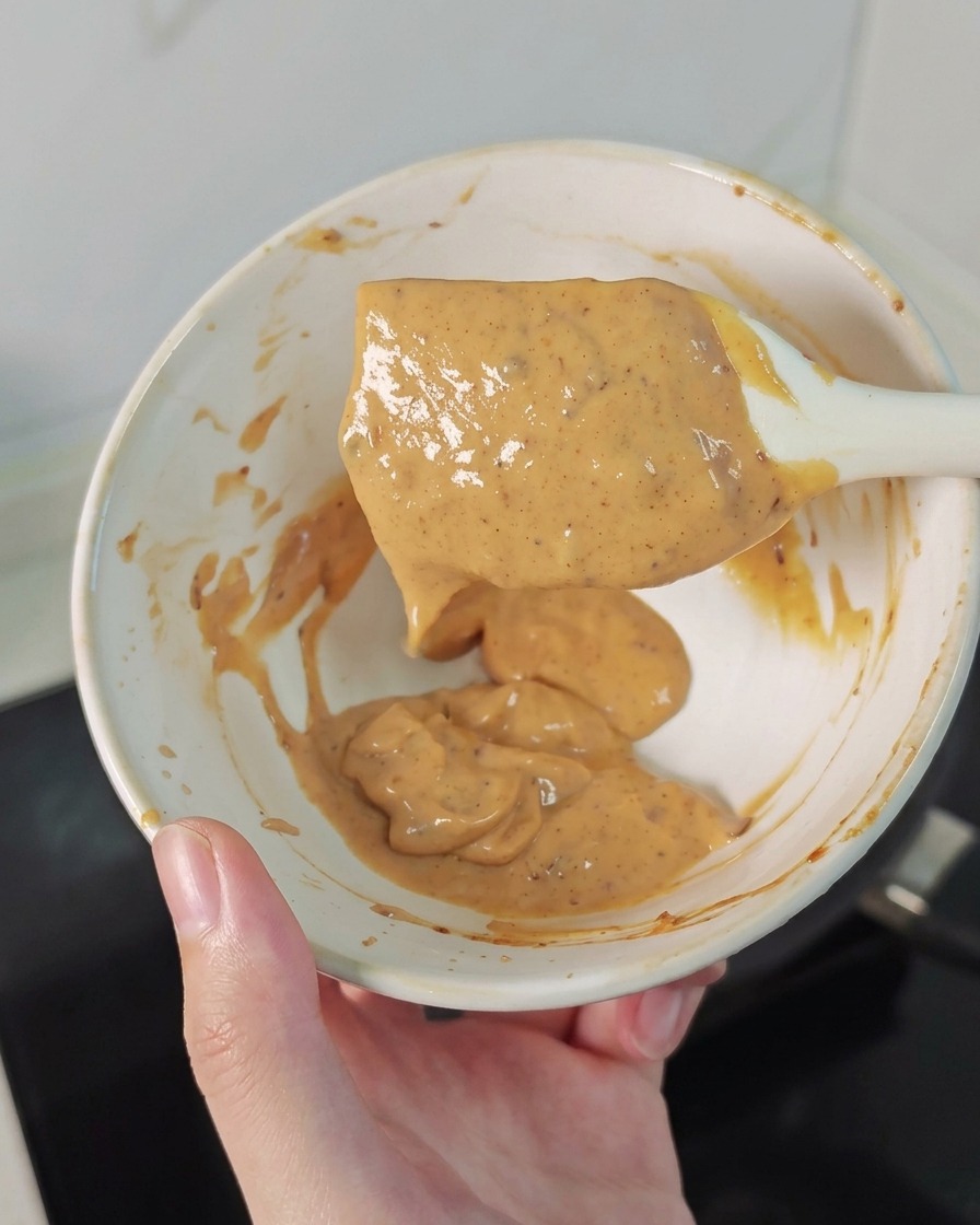 Une main tenant un bol et une spatule blanche nappée dune sauce crémeuse orangée.