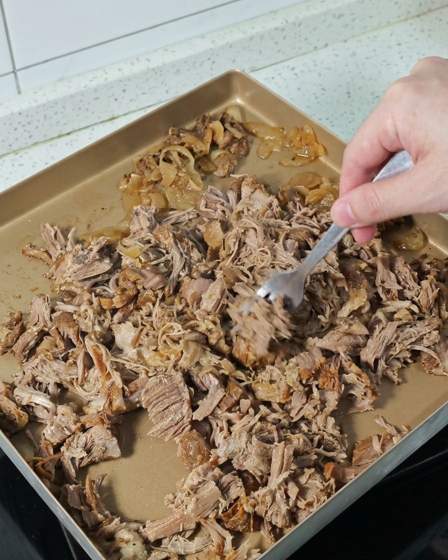 Une main utilisant une fourchette pour séparer et effilocher le porc cuit sur une plaque dorée.