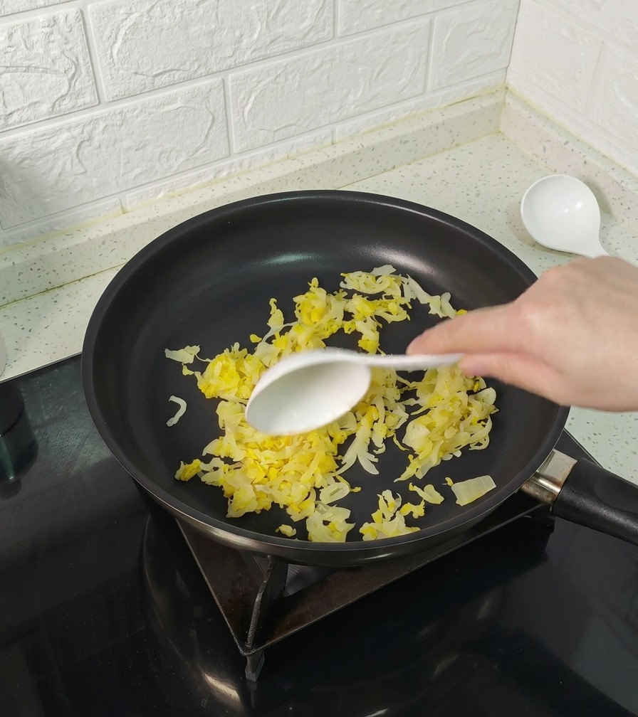 Une main utilisant une cuillère blanche pour remuer la choucroute jaune dans une poêle antiadhésive.