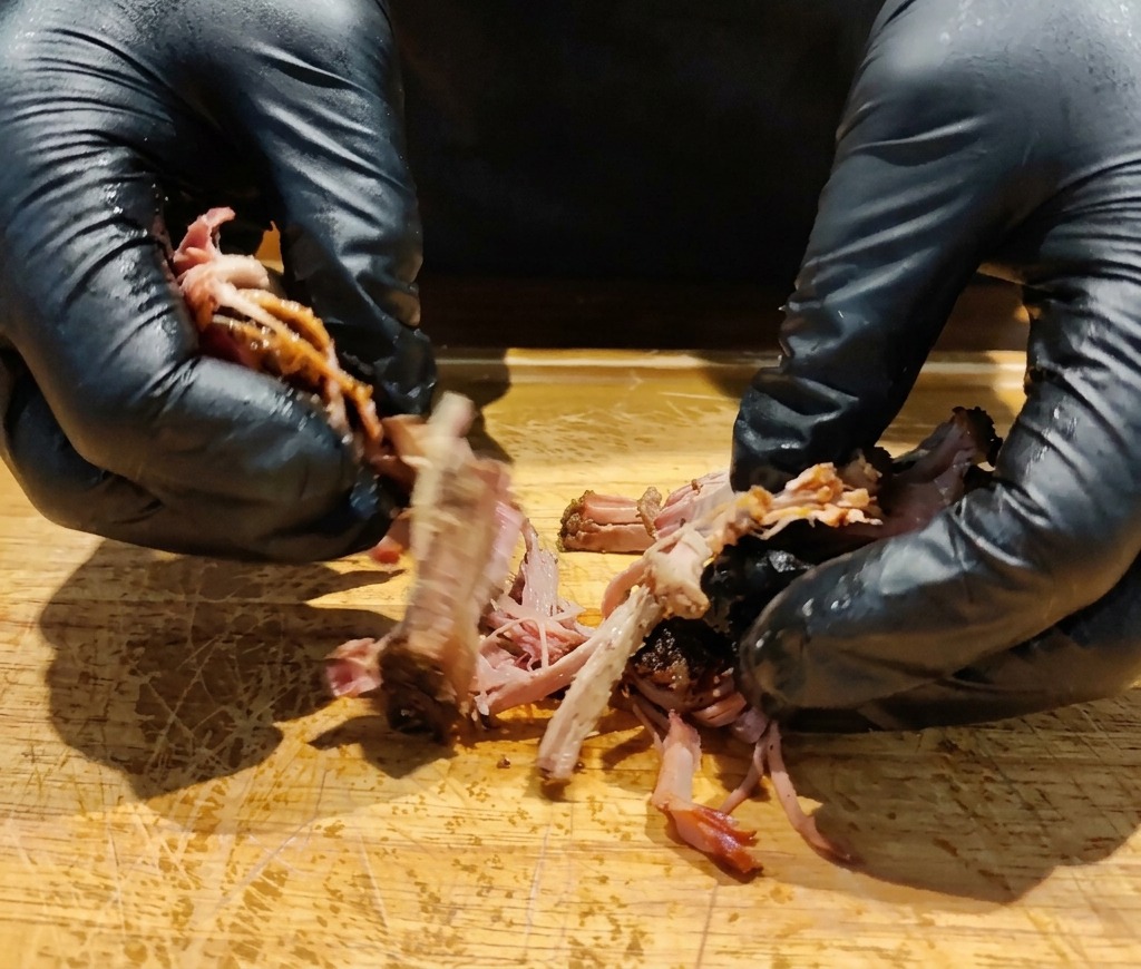 Mains gantées effilochant un morceau déchine de porc rôtie sur une planche en bois.