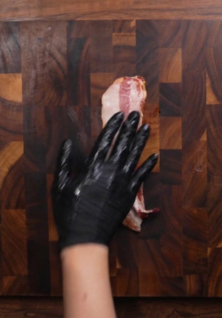 Une main gantée posée sur une tranche de bacon cru sur une planche à découper en bois sombre, sapprêtant à la trancher.