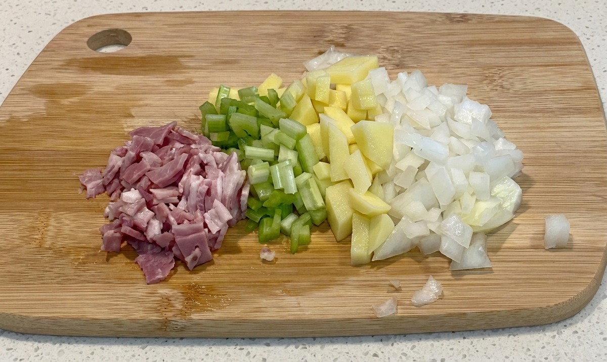 Dés de bacon, céleri, pommes de terre et oignons disposés soigneusement sur une planche à découper en bois.