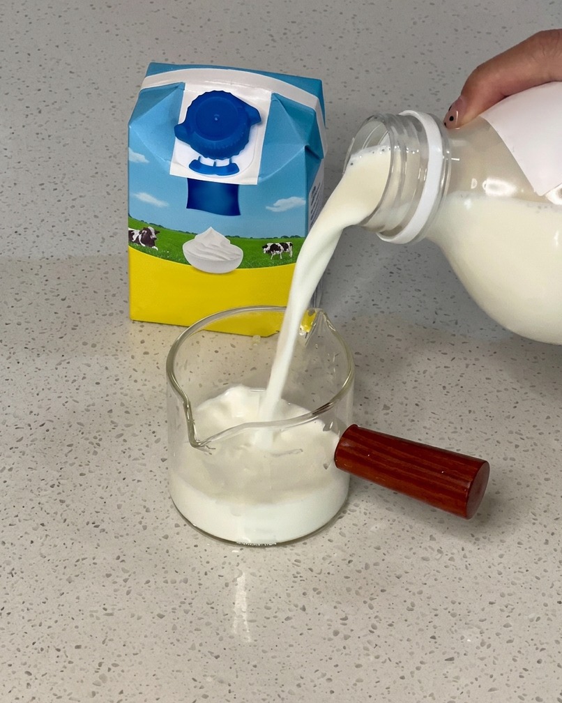 Versement de lait depuis une brique bleue et jaune dans un pichet en verre contenant de la crème.