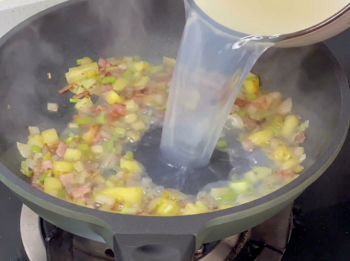 Versement du bouillon de palourdes clair dans une poêle contenant les légumes et le bacon saisis.