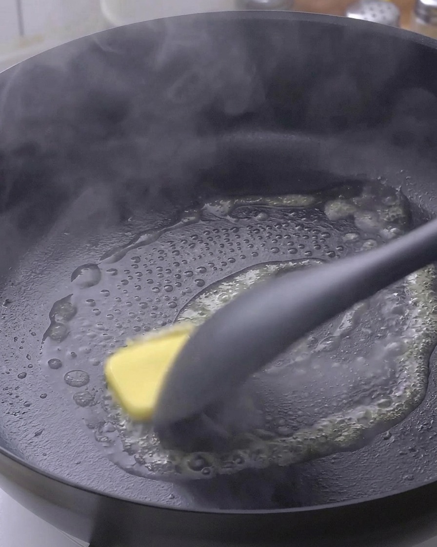 Un morceau de beurre fondant dans une poêle noire chaude, remué par une spatule.