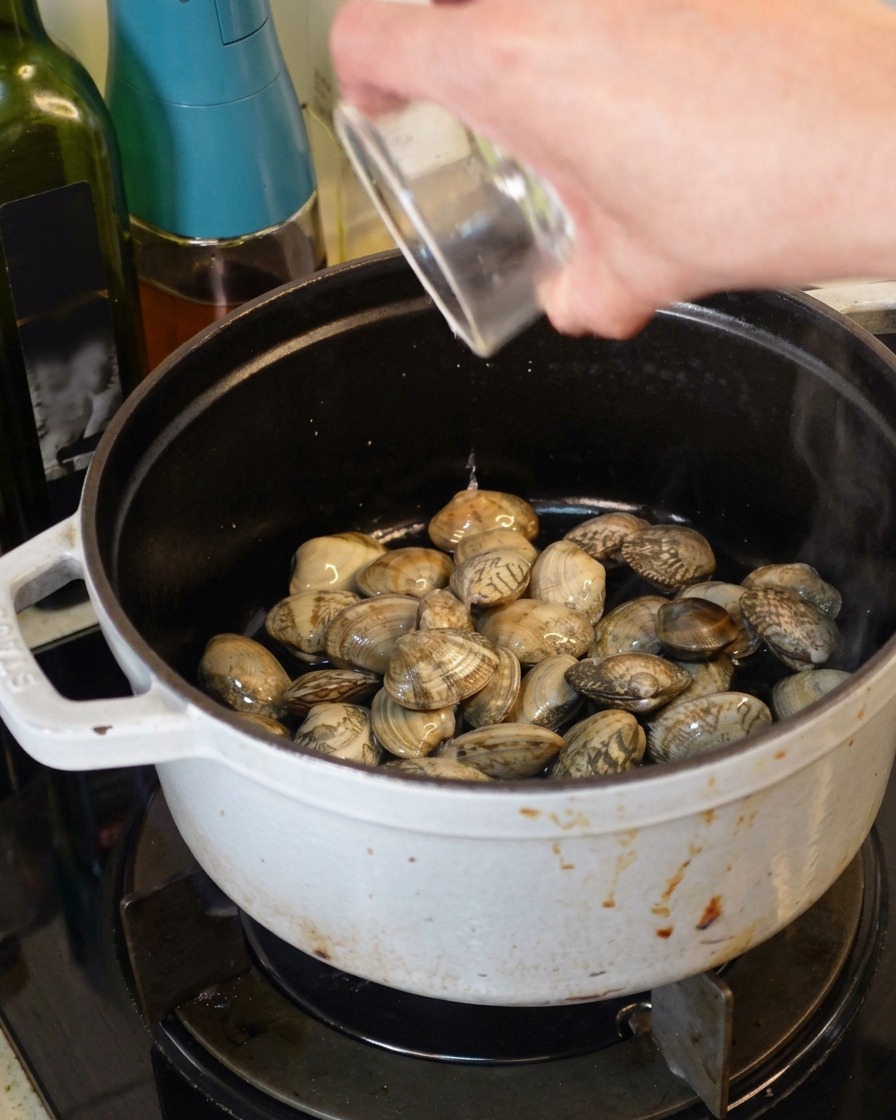 Vin blanc clair versé dun petit récipient en verre dans une cocotte en fonte blanche remplie de palourdes fraîches.