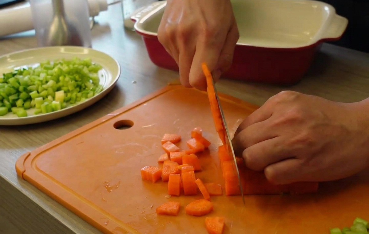 Mains utilisant un couteau de cuisine pour couper des carottes orange vif en petits cubes sur une planche à découper orange, avec du céleri haché en arrière-plan.