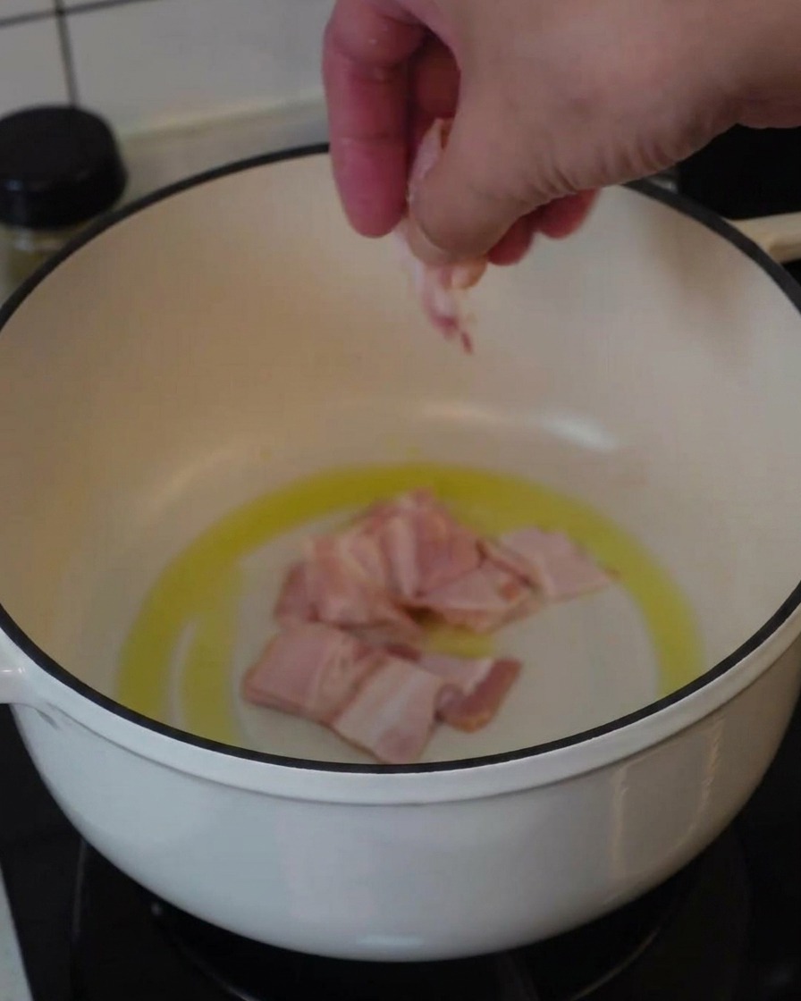 Une main plaçant des morceaux de lard cru dans une cocotte blanche avec un peu de matière grasse pour commencer la cuisson.