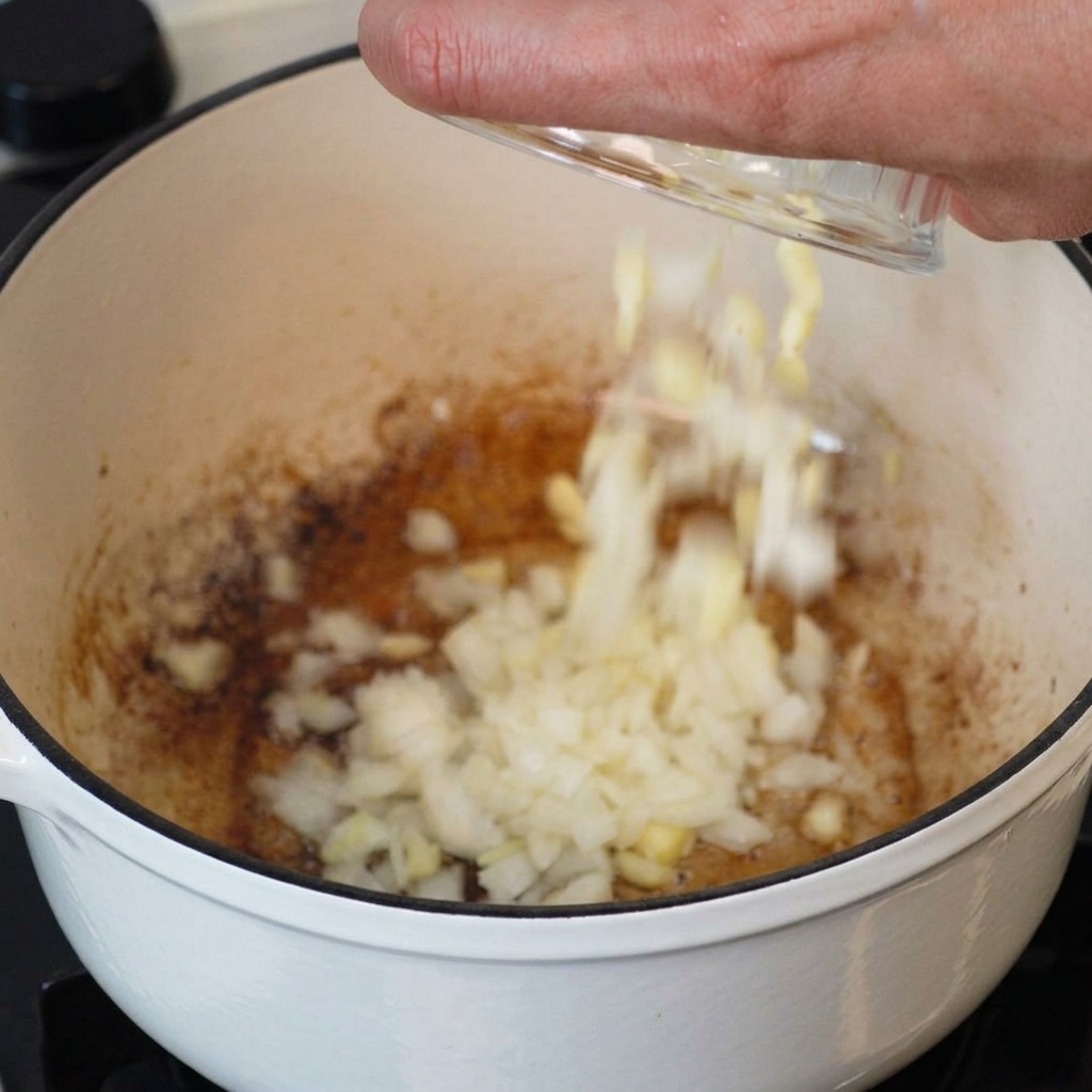 Une main versant un bol doignons hachés et dail émincé dans une cocotte blanche contenant le gras de bacon fondu.