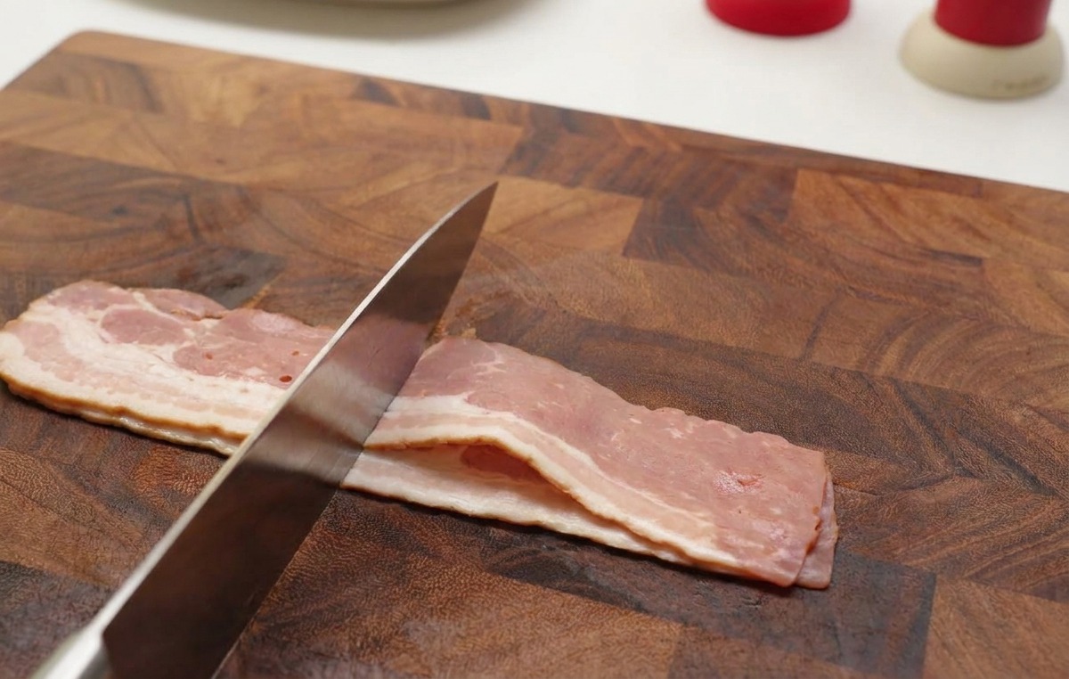 Mains utilisant un couteau tranchant pour couper des tranches de bacon cru en petits morceaux sur une planche à découper en bois.