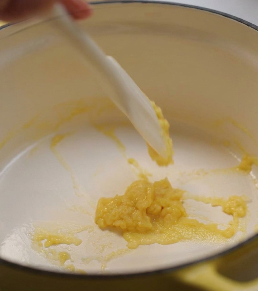Une spatule en silicone blanche remuant une pâte épaisse et jaune pâle de beurre et de farine dans une marmite jaune.