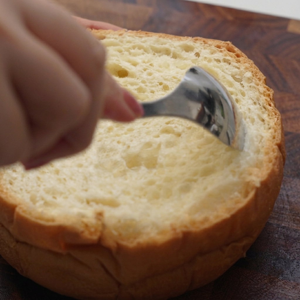 Une main utilisant une cuillère en métal pour évider lintérieur moelleux dune miche de pain ronde.