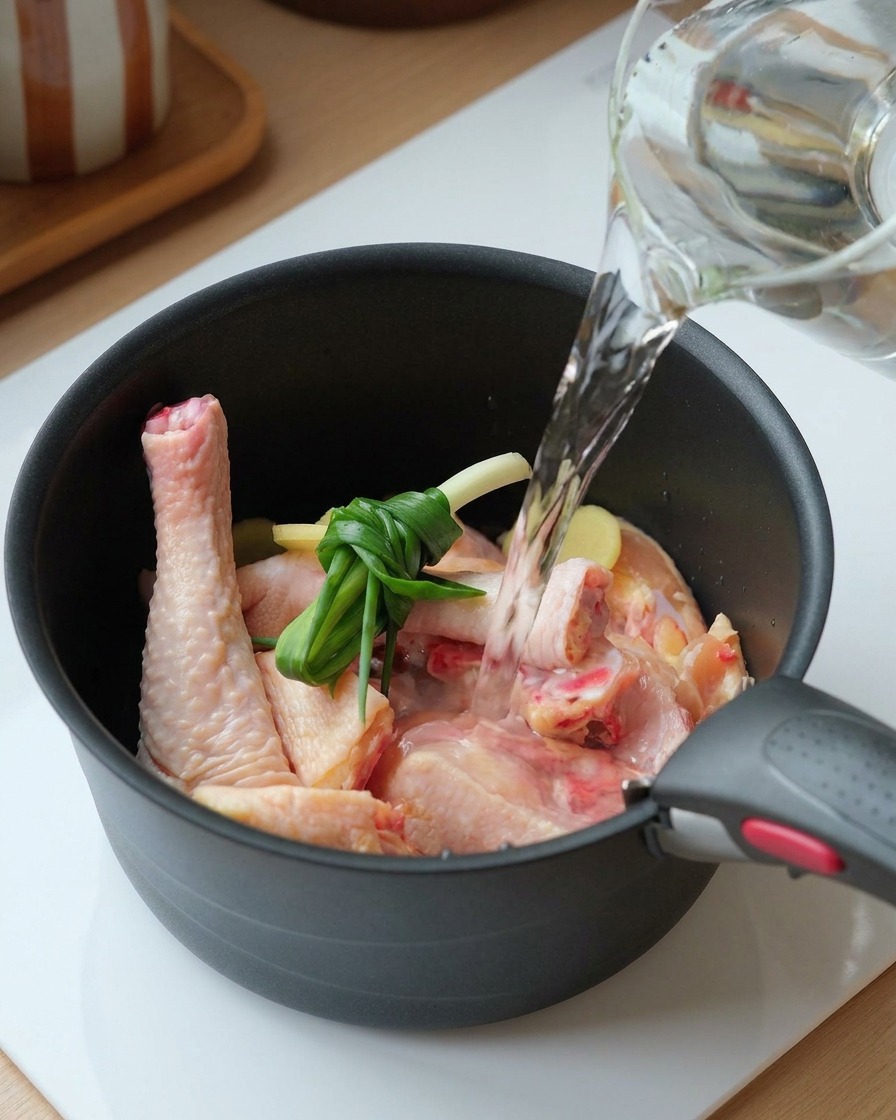 De leau claire versée dune carafe dans une marmite contenant du poulet, un bouquet doignons verts et du gingembre tranché.