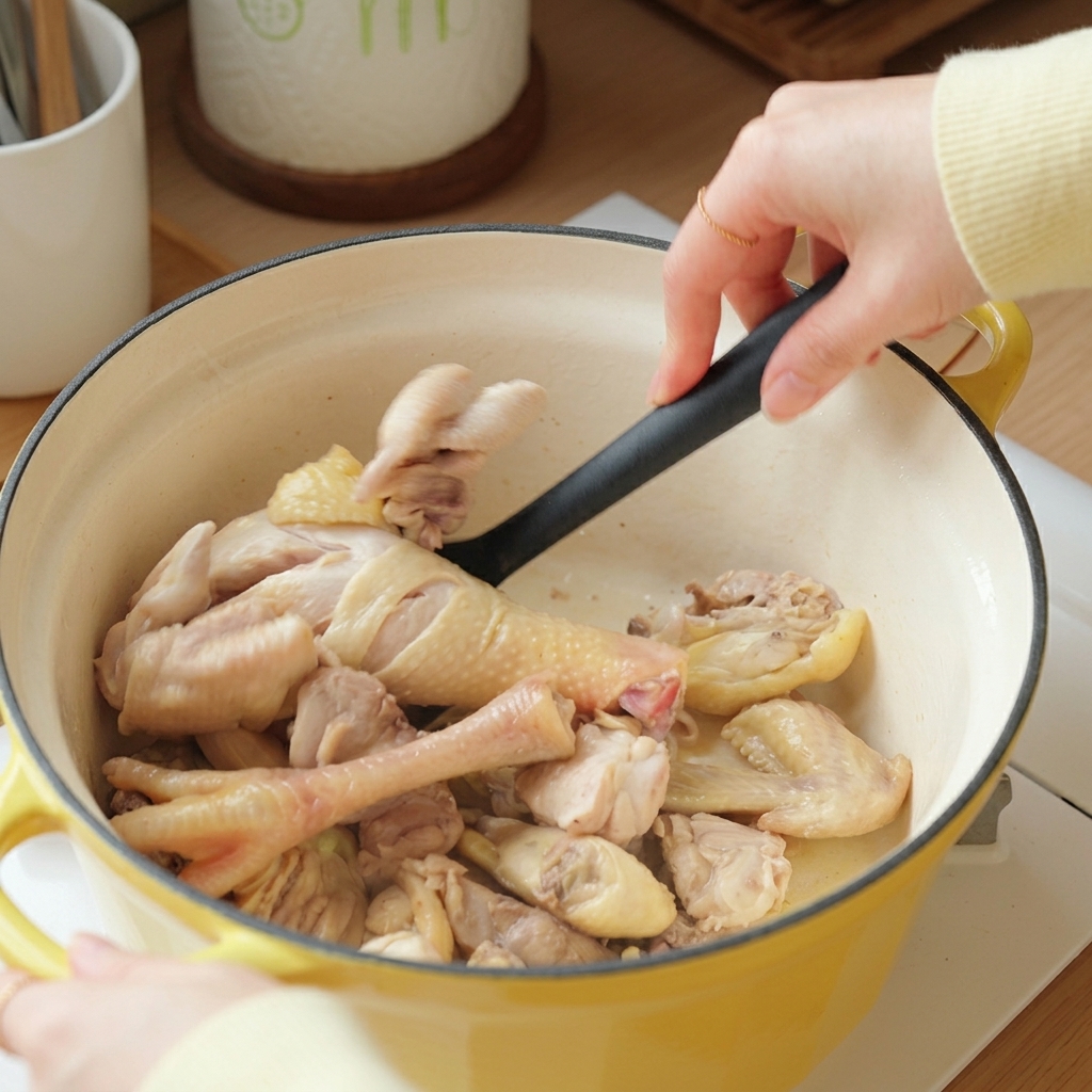 Une spatule remuant les morceaux de poulet blanchis dans la cocotte en fonte jaune.