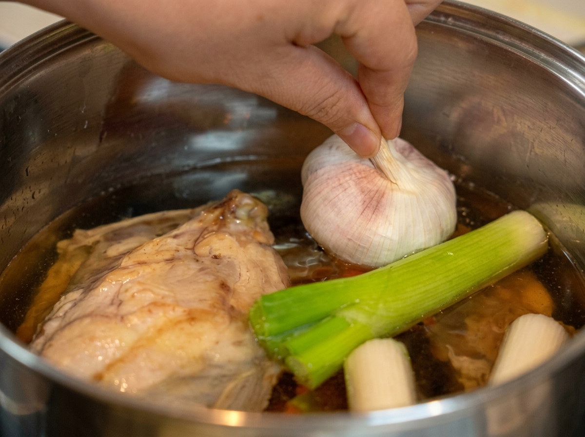 Une main déposant une tête dail entière non épluchée dans une marmite avec du poulet et des feuilles de poireau.