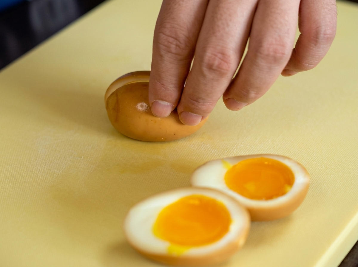 Main tranchant un œuf mollet mariné en deux sur une planche à découper, révélant un jaune orangé coulant.