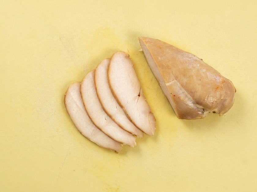 Tranches fines et régulières de blanc de poulet cuit disposées sur une planche à découper jaune.