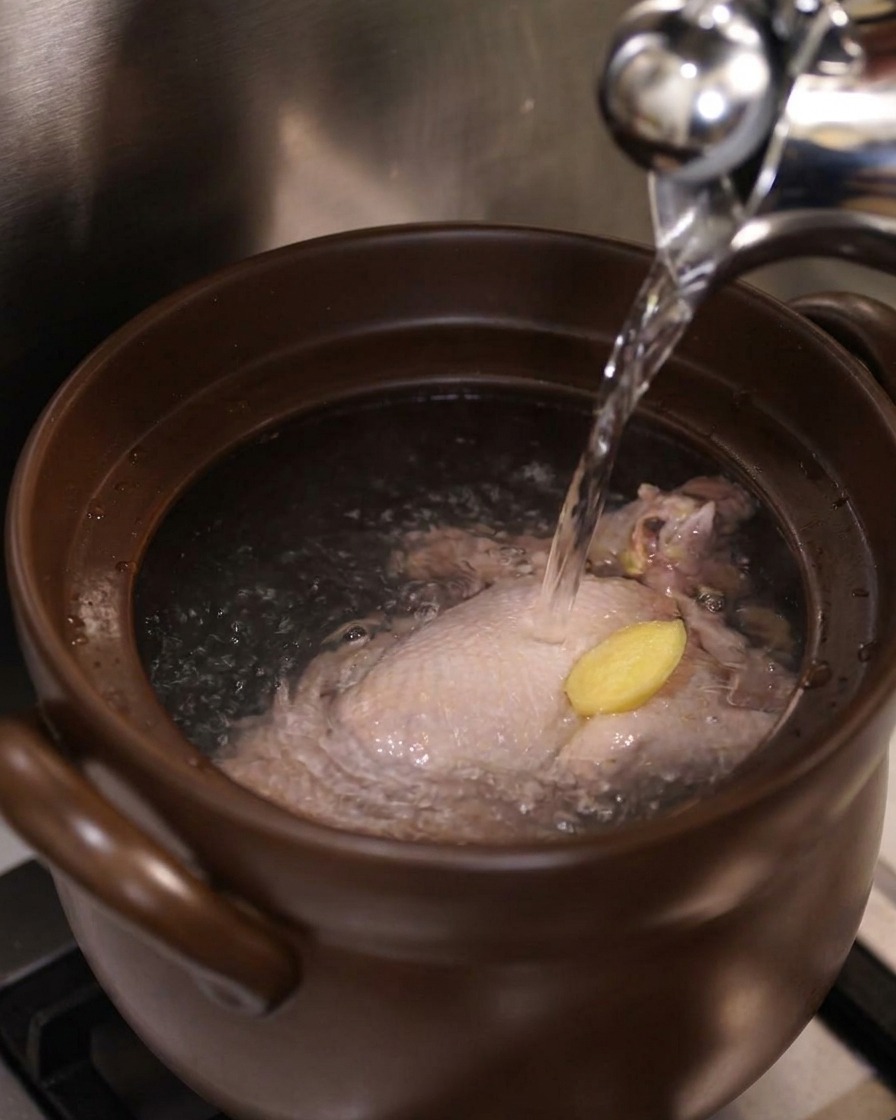 De leau bouillante versée dune bouilloire sur un poulet entier et une tranche de gingembre dans un pot.