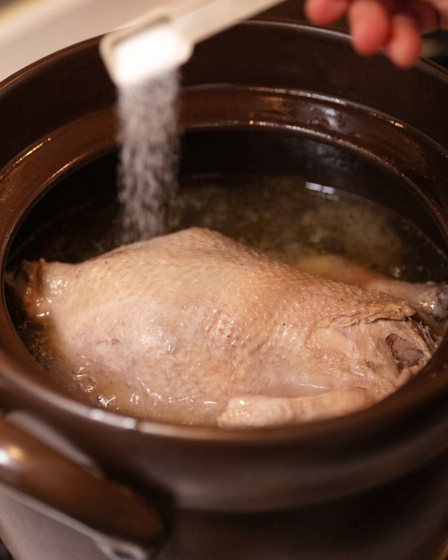 Une main utilisant une cuillère doseuse pour saupoudrer du sel sur le poulet dans le bouillon clair.