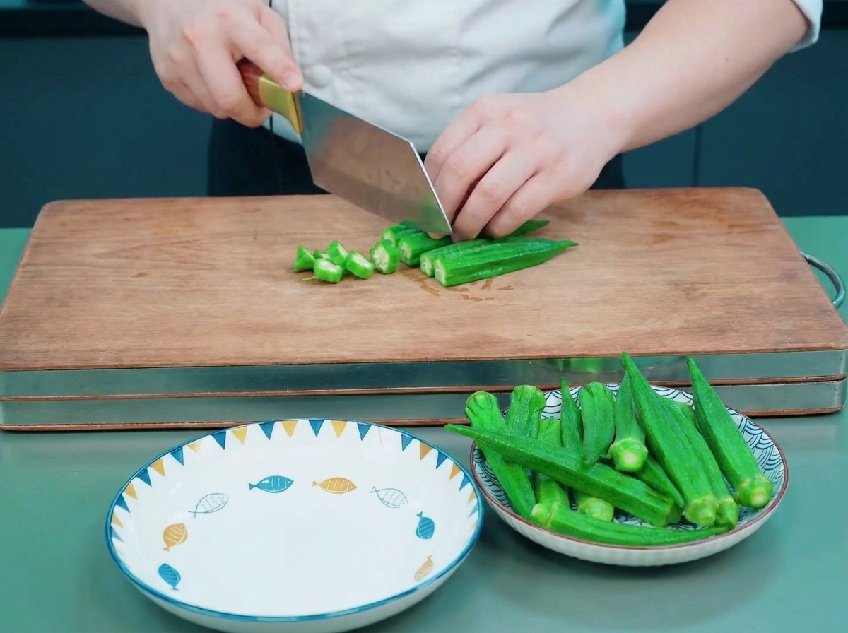 Un chef utilisant un couperet pour trancher des gombos blanchis en rondelles étoilées sur une planche en bois.