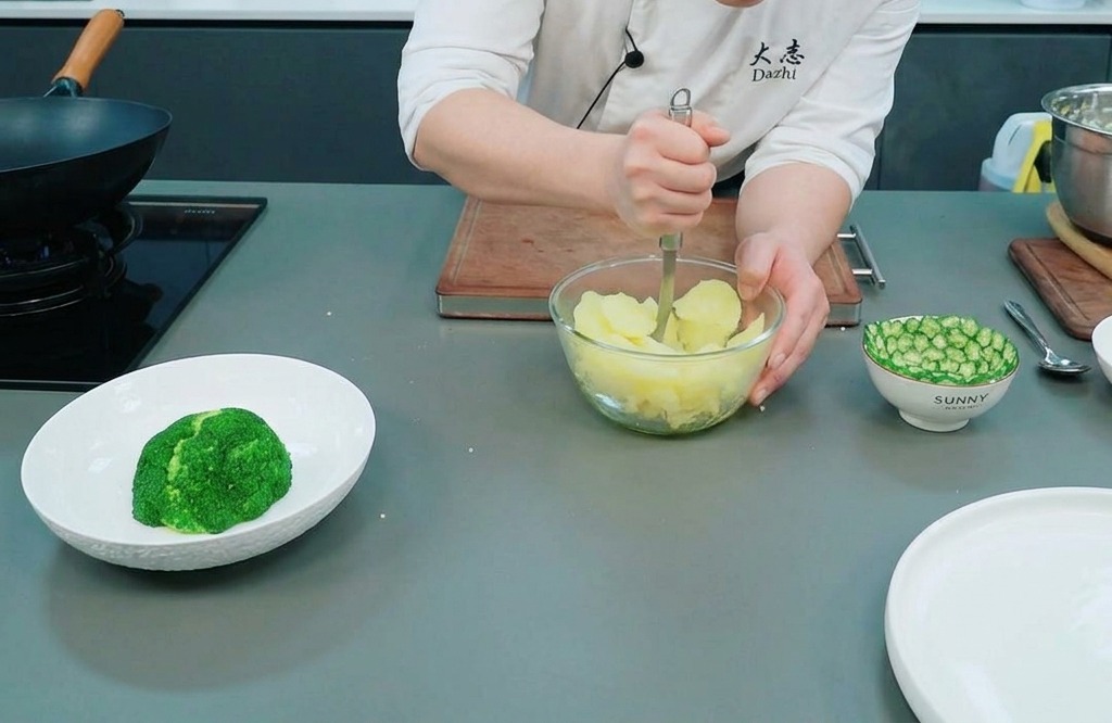Chef utilisant un presse-purée en métal pour écraser des pommes de terre cuites à la vapeur dans un bol en verre.