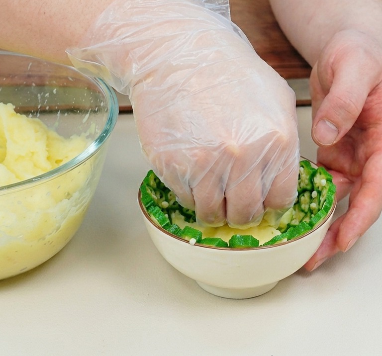 Une main gantée pressant de la purée de pommes de terre dans un bol tapissé de tranches de gombo.