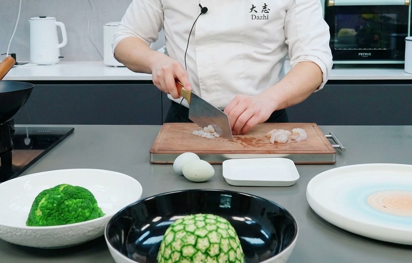 Un chef utilisant un couperet pour hacher des crevettes crues en petits morceaux sur une planche en bois.