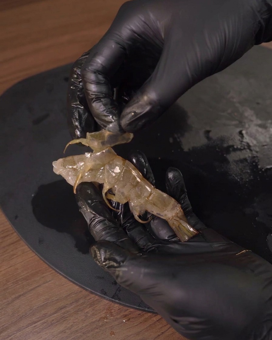 Une personne avec des gants noirs retirant la carapace translucide dune crevette crue.
