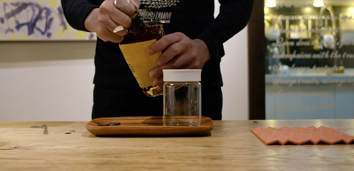 Main versant du bourbon ambré dune bouteille vers un bocal en verre transparent.