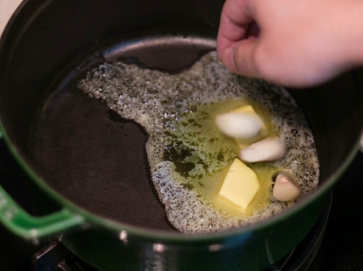 Main déposant des gousses dail dans du beurre moussant à lintérieur dune cocotte verte.