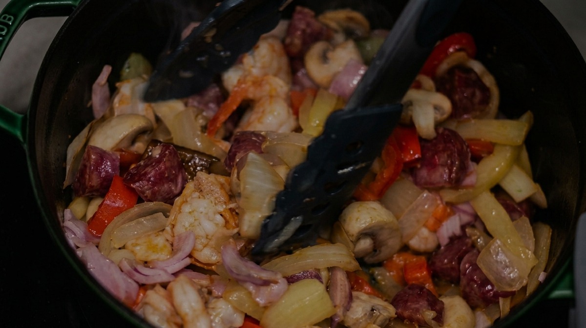 Mélange de saucisse fumée, bacon et crevettes remué avec des légumes à laide dune pince.