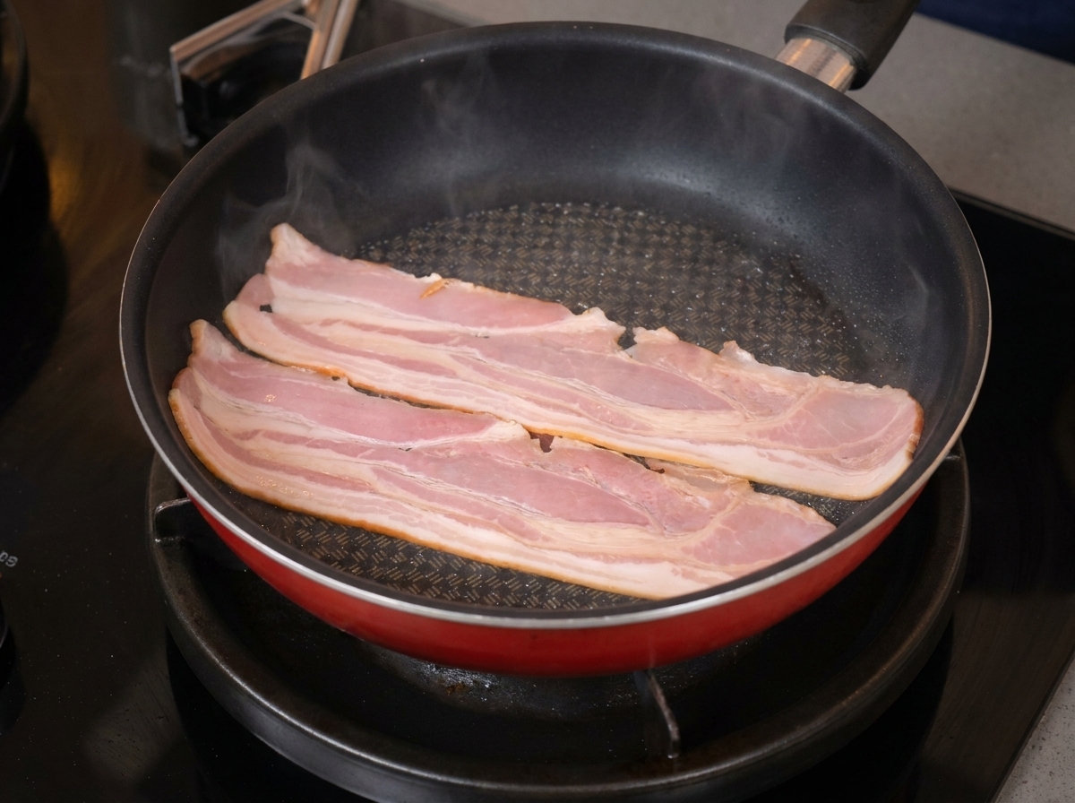 Deux tranches de bacon qui grésillent et commencent à se courber dans une poêle rouge.