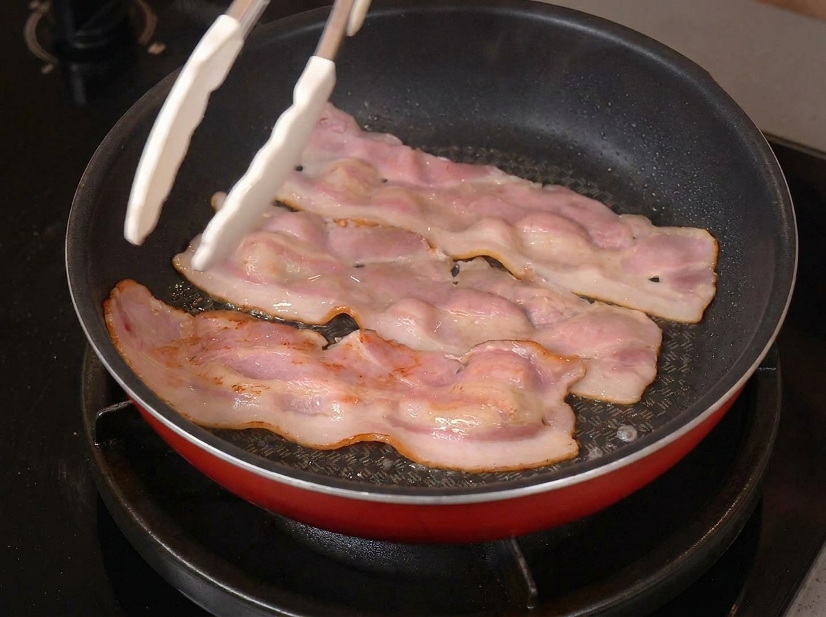Trois tranches de bacon dans une poêle, les bords brunis, avec une pince de cuisine blanche.