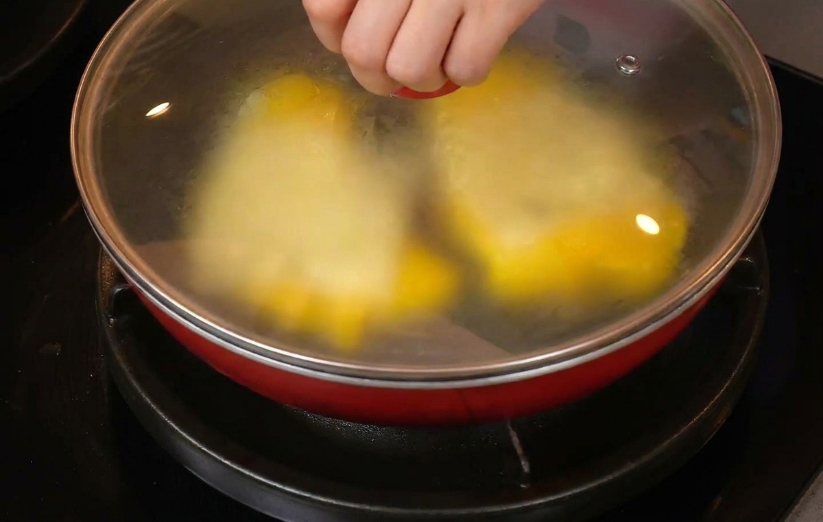 Une main posant un couvercle en verre sur une poêle contenant des œufs et du fromage.