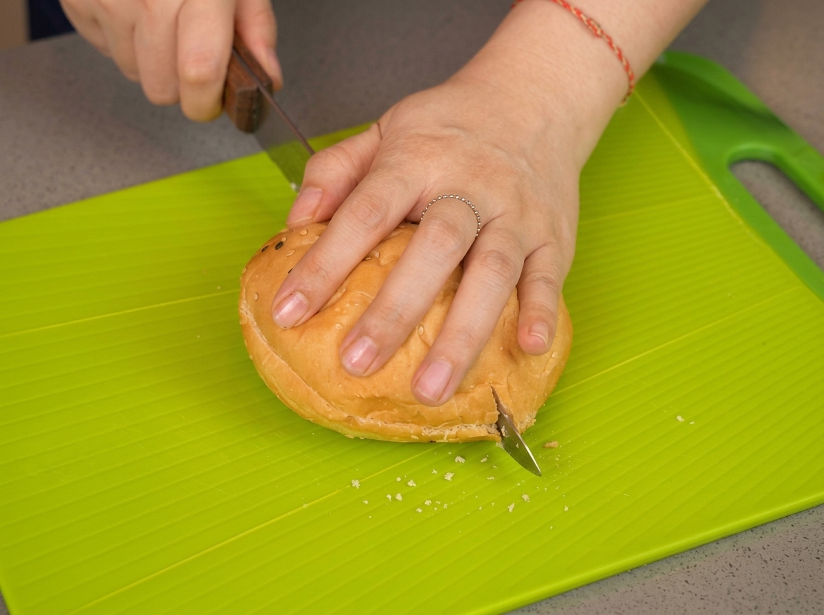 Main pressant un pain burger pendant quun couteau scie le milieu sur une planche verte.