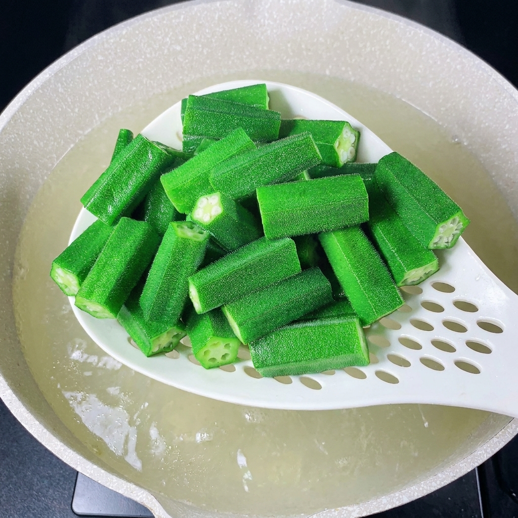 Une écumoire blanche retirant des morceaux de gombos fraîchement blanchis de leau bouillante.