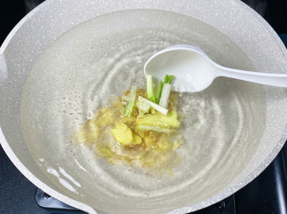 Tranches de gingembre frais et segments de ciboule ajoutés dans leau bouillante avec une louche blanche.