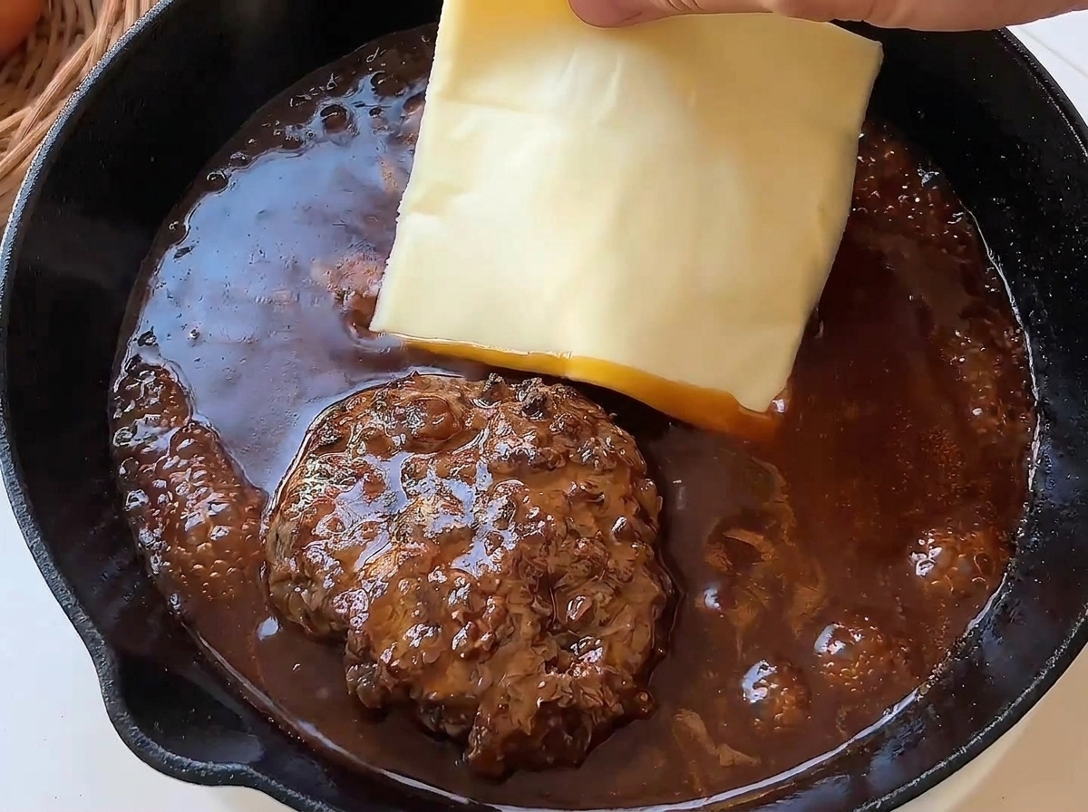 Une main déposant une tranche de fromage jaune sur un steak haché fumant.