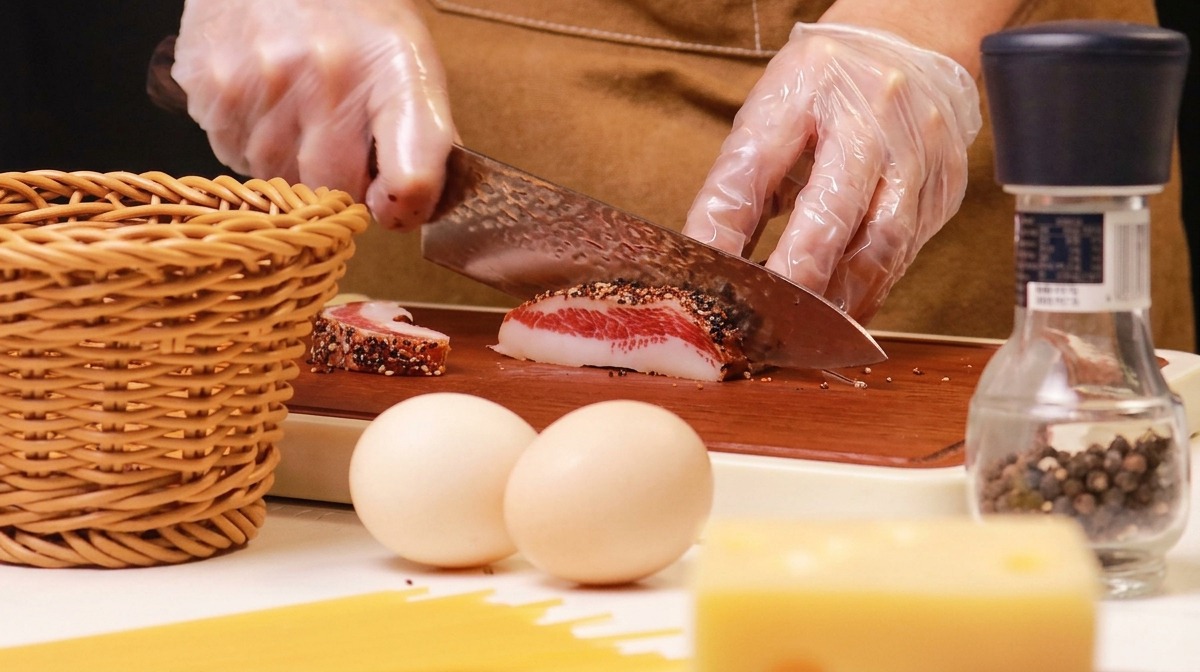 Mains gantées coupant une pièce de guanciale poivré sur une planche en bois.