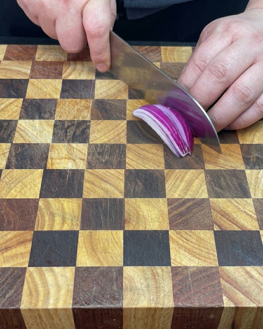 Une main tenant un couteau pour émincer un oignon rouge sur une planche à découper.