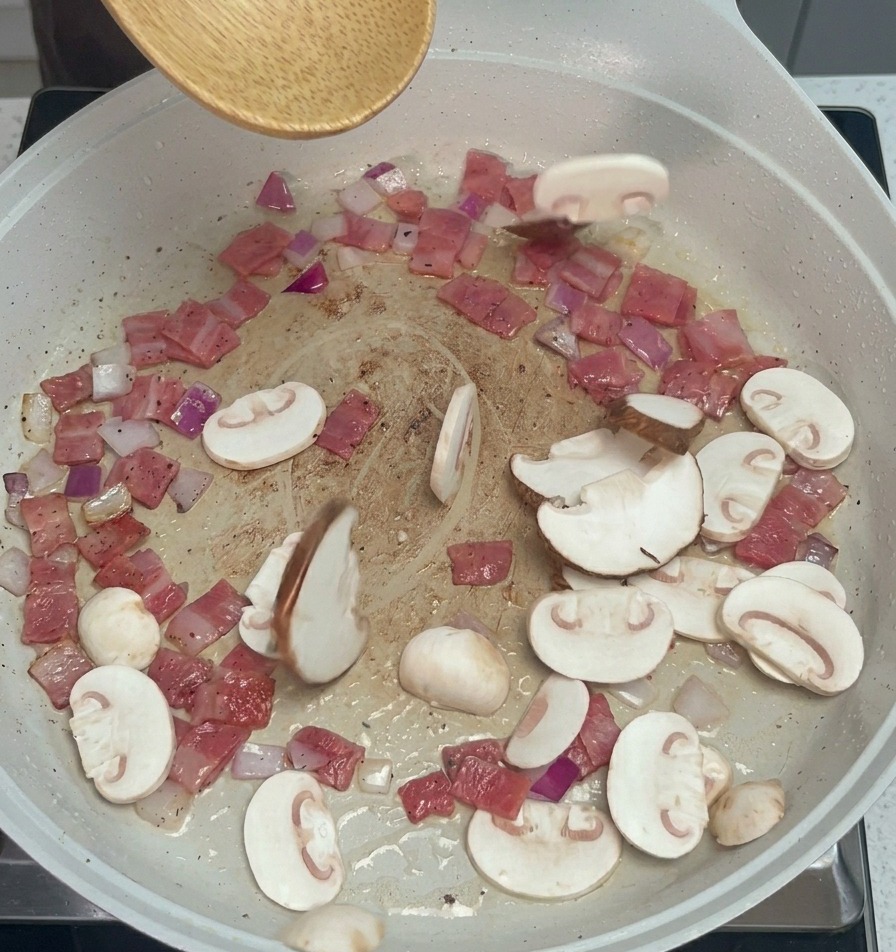 Lamelles de champignons ajoutées à la poêle avec le bacon et les oignons à laide dune cuillère en bois.