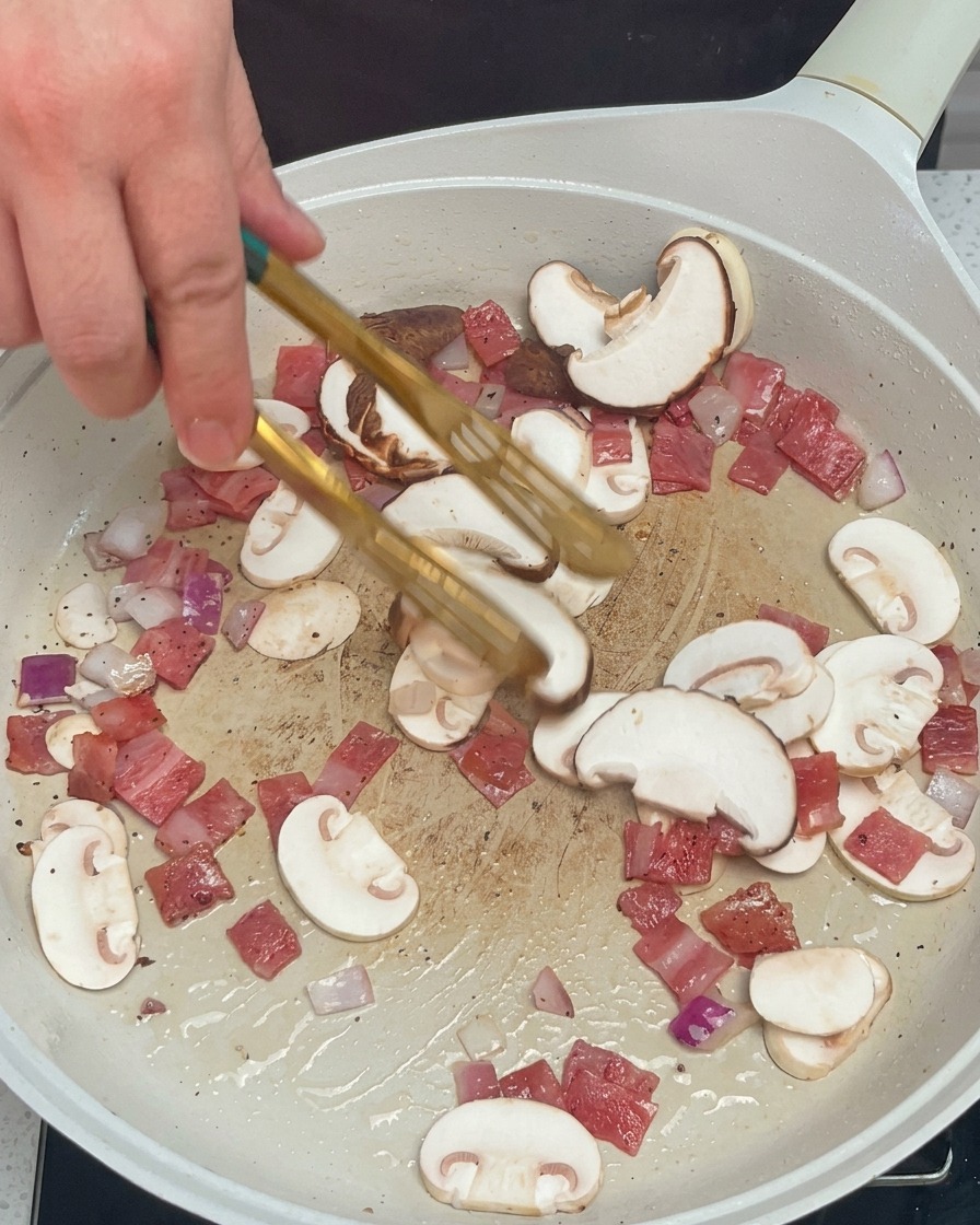 Mélange de champignons, oignons et bacon remué avec une pince dans la poêle blanche.
