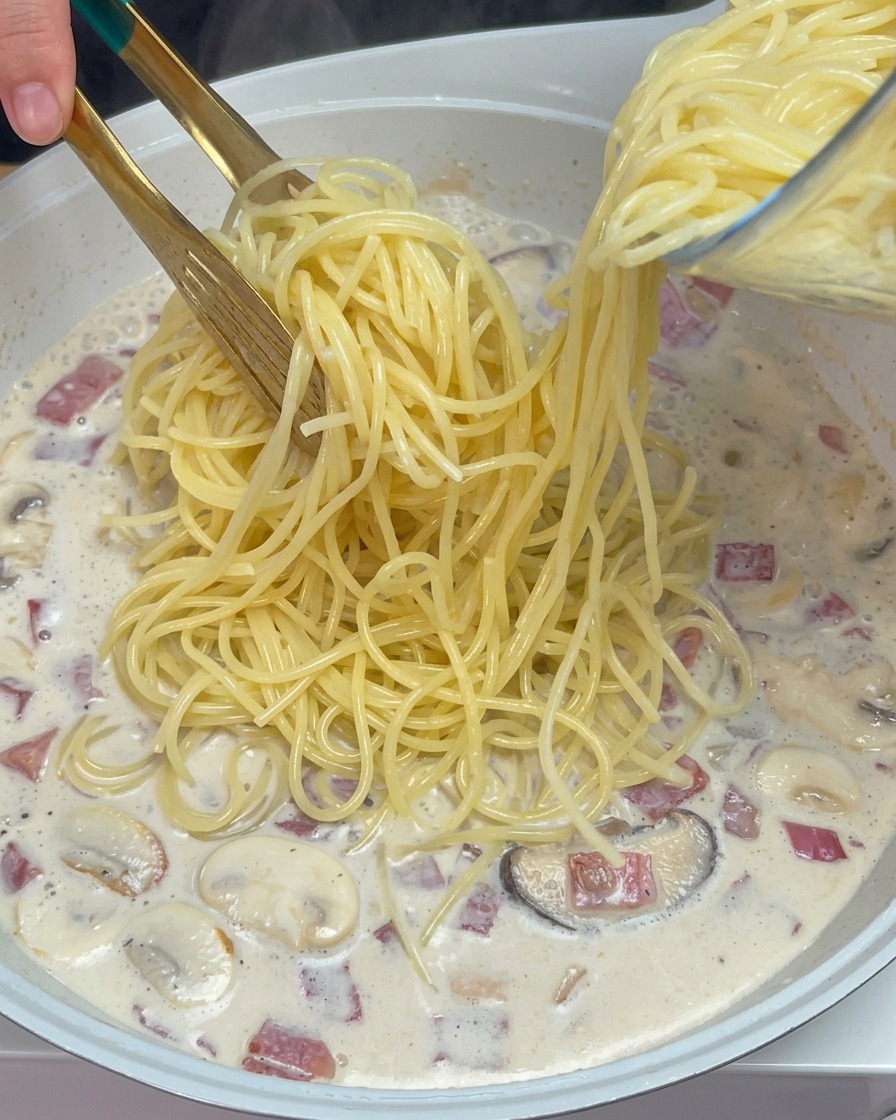 Pince déposant les spaghetti cuits dans la sauce crémeuse aux champignons et bacon.