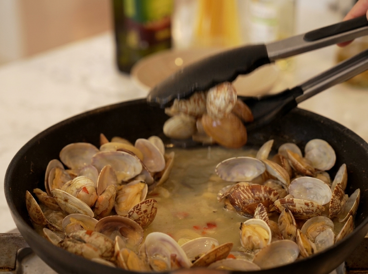 Une pince de cuisine prélevant des palourdes ouvertes dune sauteuse frémissante.